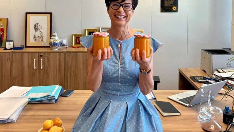 A smiling Marta Kos sitting on the edge of an office desk, holding up two jars of homemade orange jam. A small bowl of fresh apricots rests on the desk next to her, with office documents, a laptop, and framed pictures visible in the background.