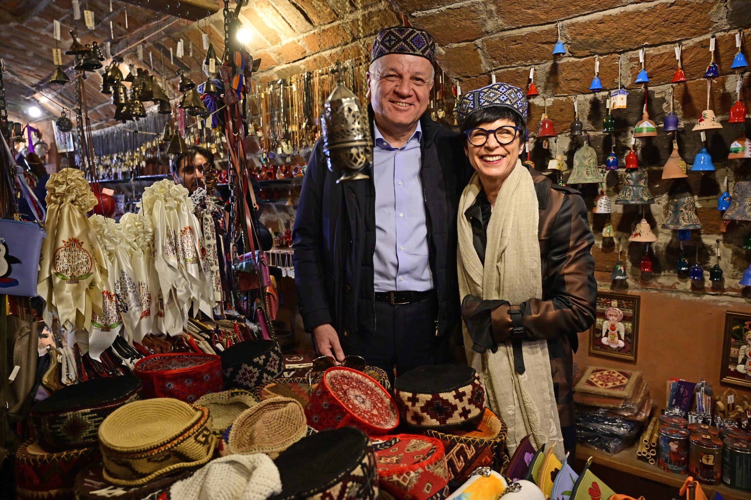 EU CSSR Marta Kos and EU Ambassador to Armenia Vassilis Maragos wearing traditional Armenian hats while visiting a local craft shop filled with handmade bells and textiles.