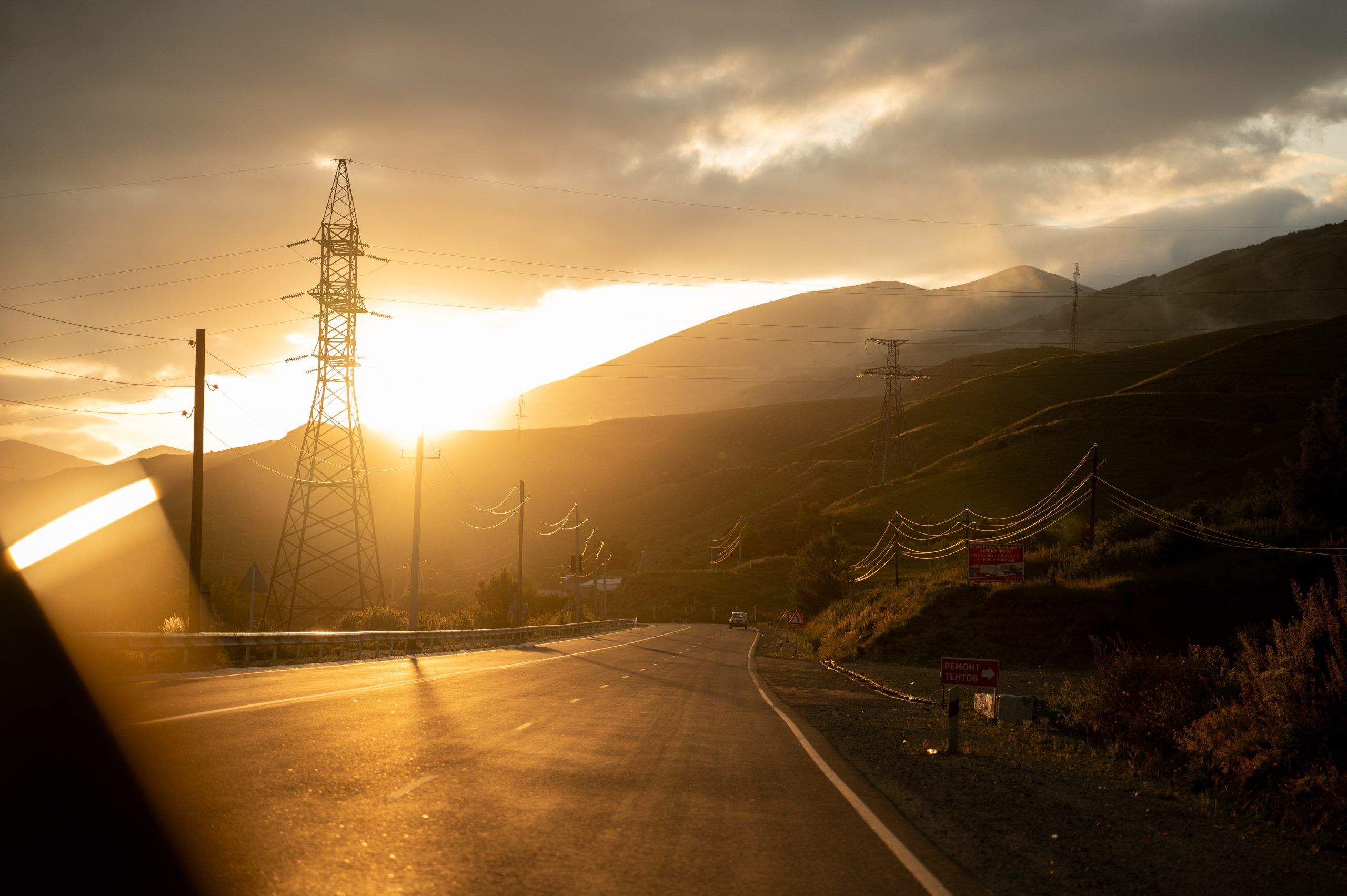 A scenic sunset over a highway in Armenia, featuring high-voltage power lines and electricity pylons stretching across rolling hills and mountains.