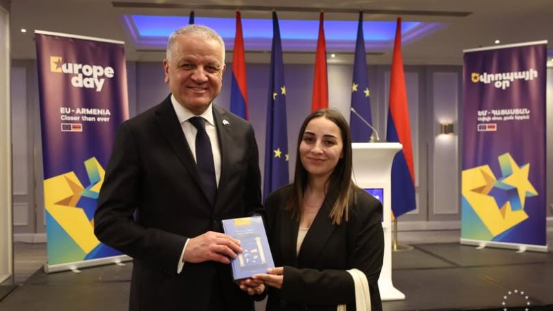 EU Ambassador to Armenia Vassilis Maragos and a young woman standing on a stage, smiling and holding a book together during a Europe Day event in Armenia.