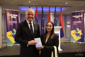 EU Ambassador to Armenia Vassilis Maragos and a young woman standing on a stage, smiling and holding a book together during a Europe Day event in Armenia.
