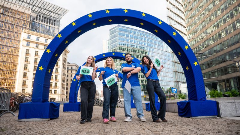 Four young people in blue shirts stand smiling under a large blue inflatable arch decorated with yellow EU stars, holding signs that read "YEA 10" in a city plaza surrounded by modern buildings.