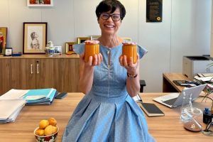 A smiling Marta Kos sitting on the edge of an office desk, holding up two jars of homemade orange jam. A small bowl of fresh apricots rests on the desk next to her, with office documents, a laptop, and framed pictures visible in the background.