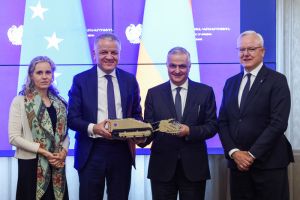 Four officials standing together indoors, posing for a photograph. In the center, EU Ambassador Vassilis Maragos and Armenian Deputy Prime Minister Mher Grigoryan smile while jointly holding a small, detailed scale model of a tan-colored, tracked demining vehicle.