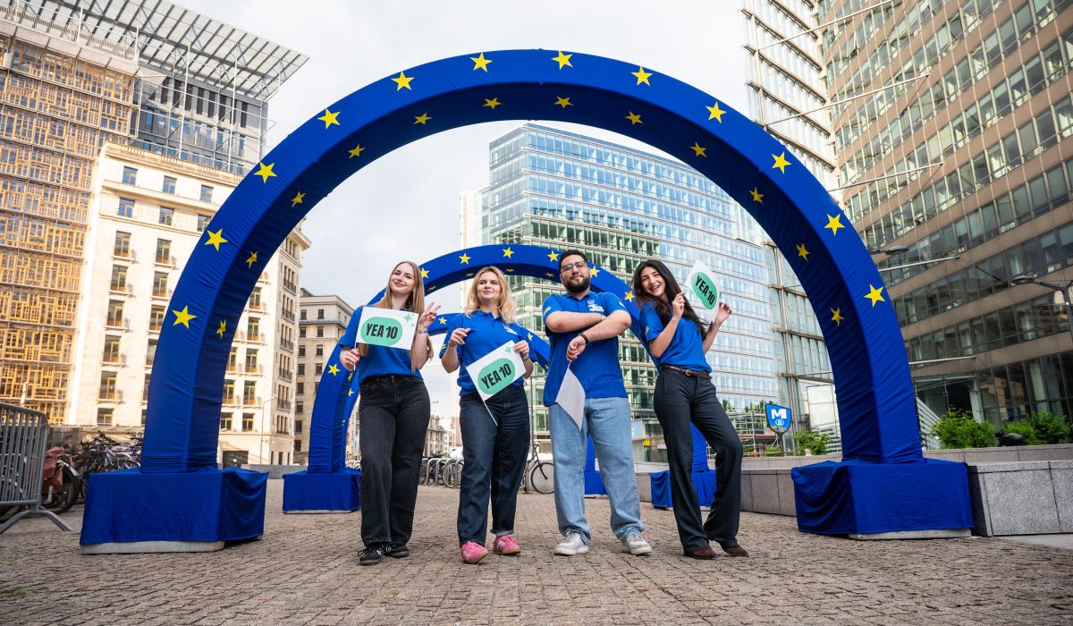 Four young people in blue shirts stand smiling under a large blue inflatable arch decorated with yellow EU stars, holding signs that read "YEA 10" in a city plaza surrounded by modern buildings.